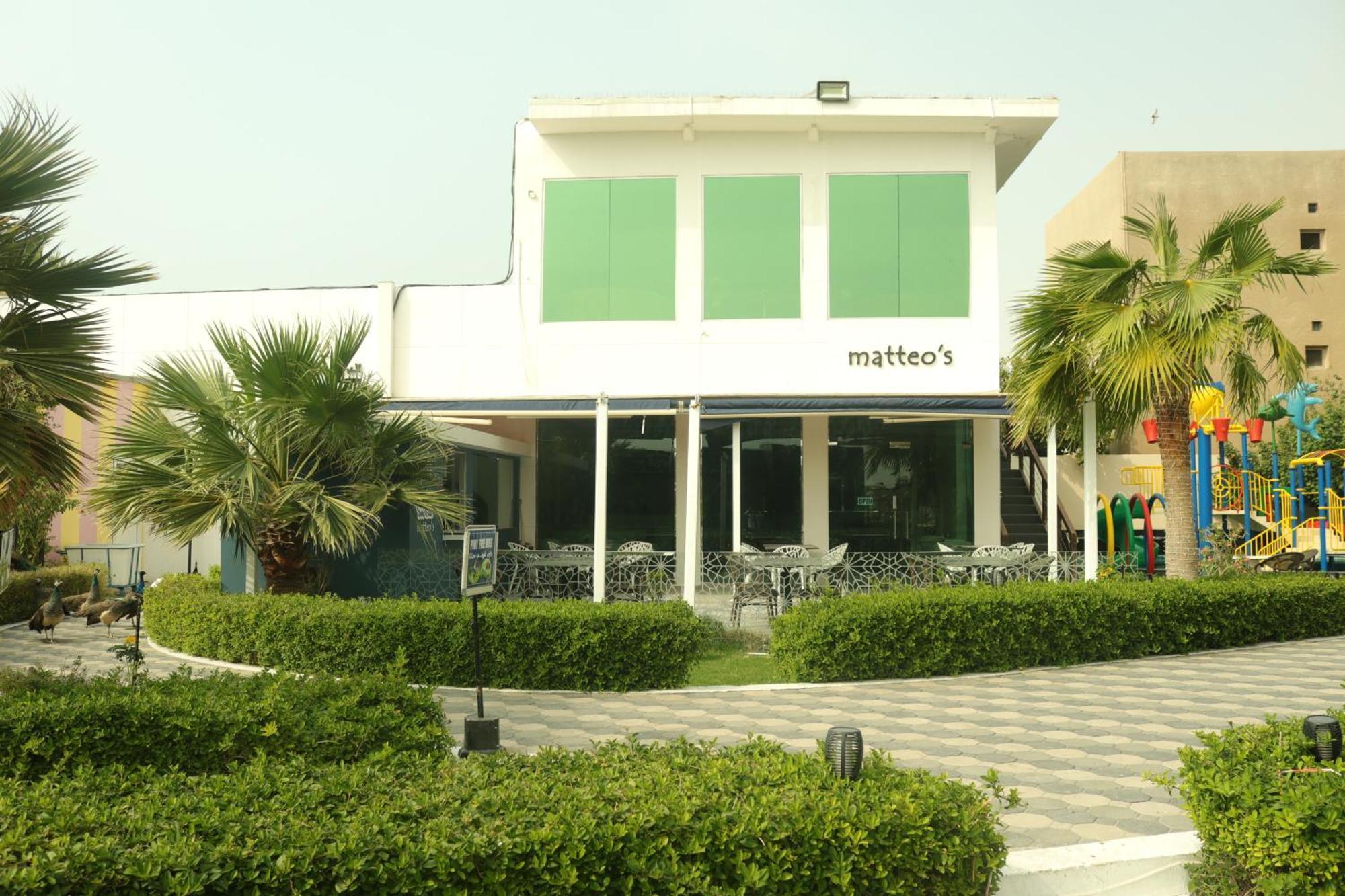 Lagoona Beach Luxury Resort And Spa Manama Exterior photo The photo shows a building with the name "matteo's" displayed prominently on the front. The structure features large windows and green shutters. There is a landscaped area in front with bushes and palm trees, along with a paved pathway leading up to 