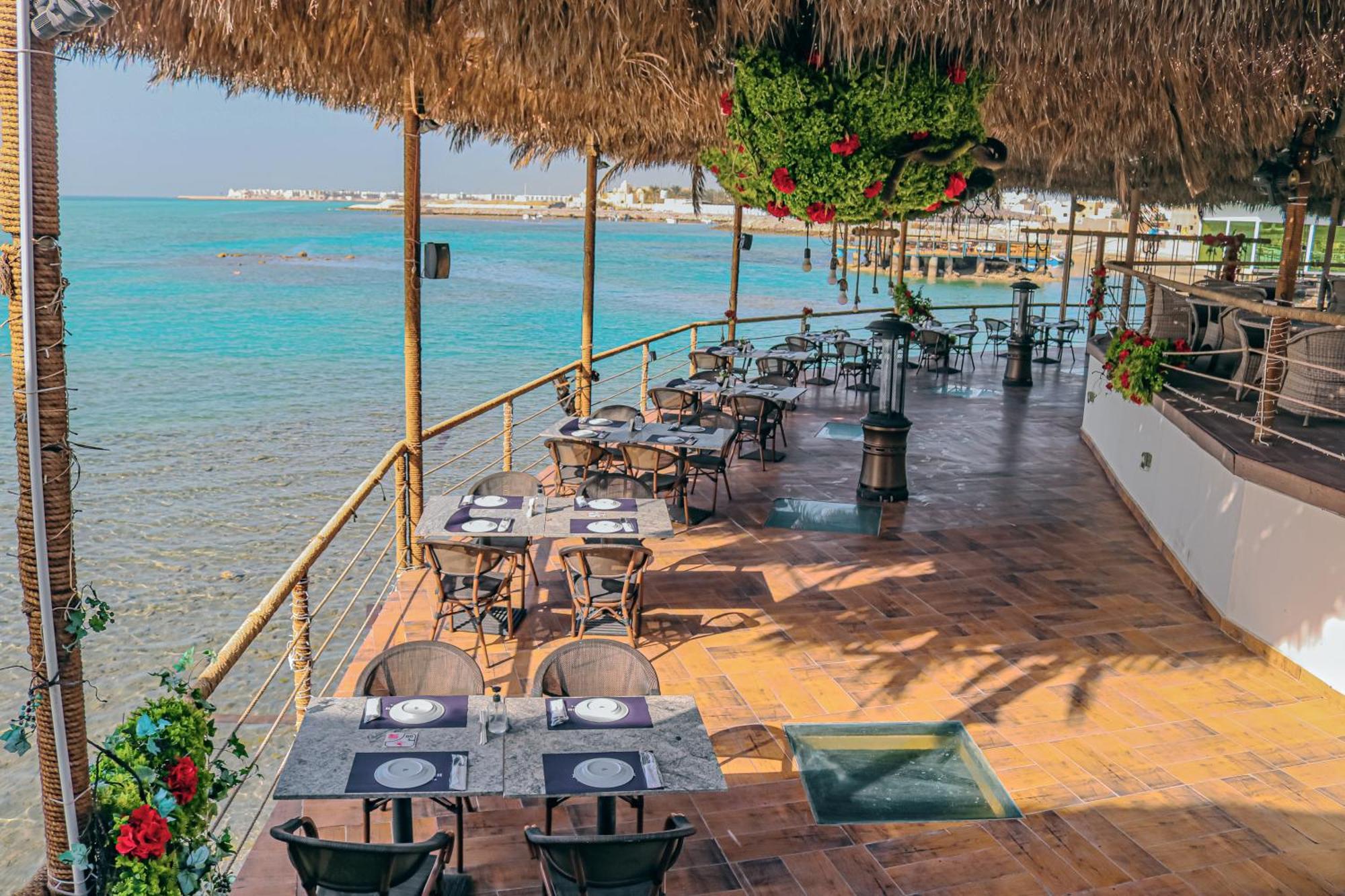 Lagoona Beach Luxury Resort And Spa Manama Exterior photo The photo shows a seaside restaurant with a spacious outdoor dining area. It features several tables set with plates and utensils, surrounded by lush greenery and decorative plants. The flooring is made of brown tiles, and there are thatched roofs pr