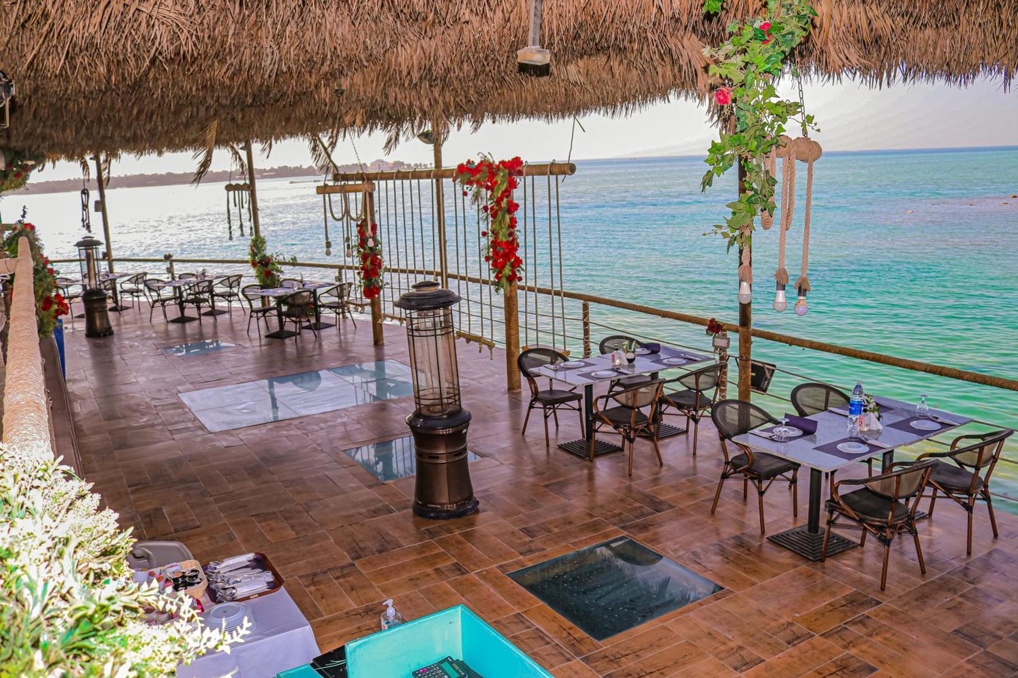 Lagoona Beach Luxury Resort And Spa Manama Exterior photo The photo depicts a coastal restaurant or dining area with an open-air design. It features a wooden floor and tables set for dining, surrounded by chairs. The space is partially covered by a thatched roof, providing shade while allowing for a view of