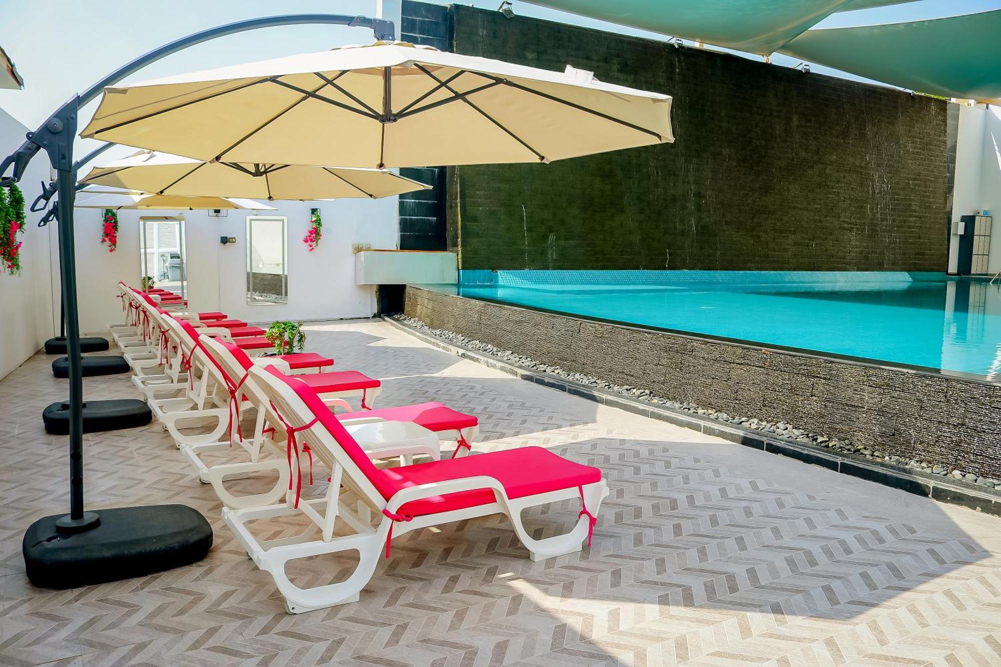 Lagoona Beach Luxury Resort And Spa Manama Exterior photo The photo shows a sunny poolside area with several lounge chairs arranged in a row. Each chair features red cushions and is positioned under large, beige umbrellas for shade. In the background, there is a beautiful swimming pool with clear blue water