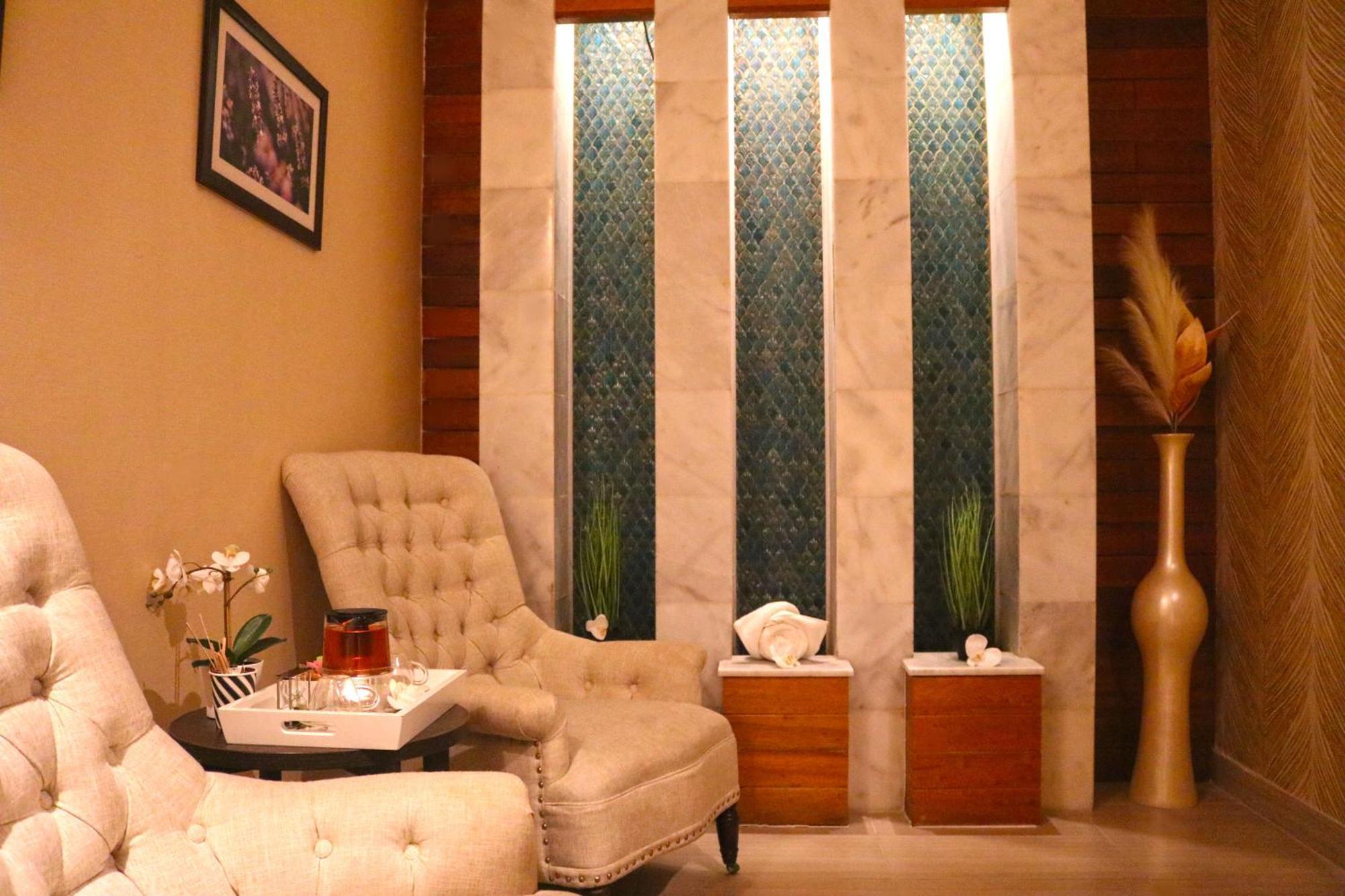 Lagoona Beach Luxury Resort And Spa Manama Exterior photo The image shows a relaxing interior space, likely a waiting area or lounge. There are two plush, beige armchairs positioned near a small table, which holds a glass pitcher and a cup. The background features tall, elegant windows with decorative blue 
