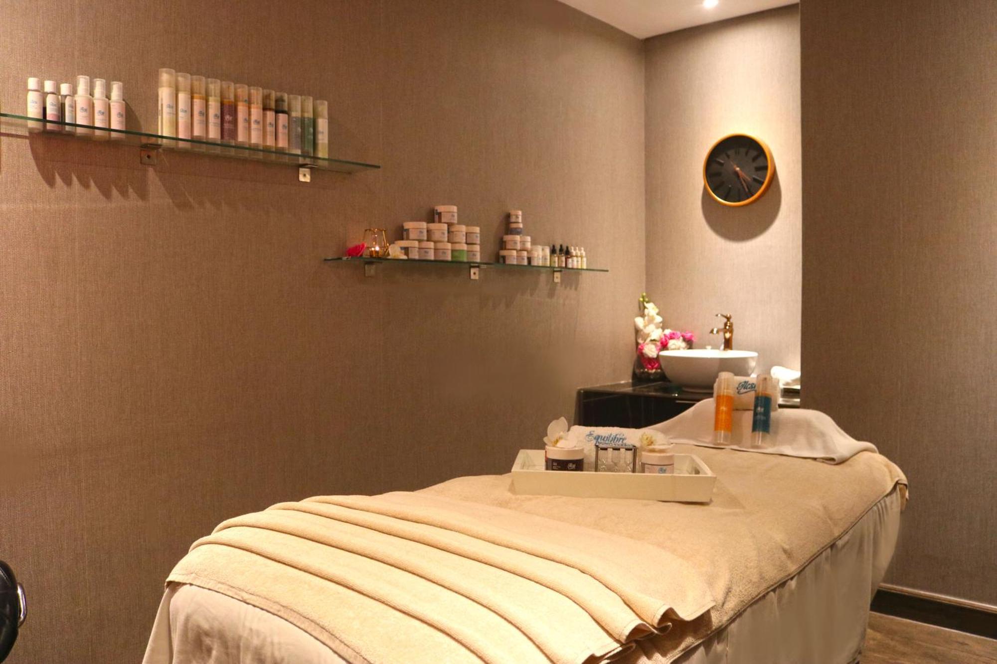 Lagoona Beach Luxury Resort And Spa Manama Exterior photo The photo shows a spa treatment room. There is a massage table draped with a light-colored blanket, creating a relaxing atmosphere. On the wall, there are shelves displaying various skincare products in bottles and jars. A clock is mounted on the wal
