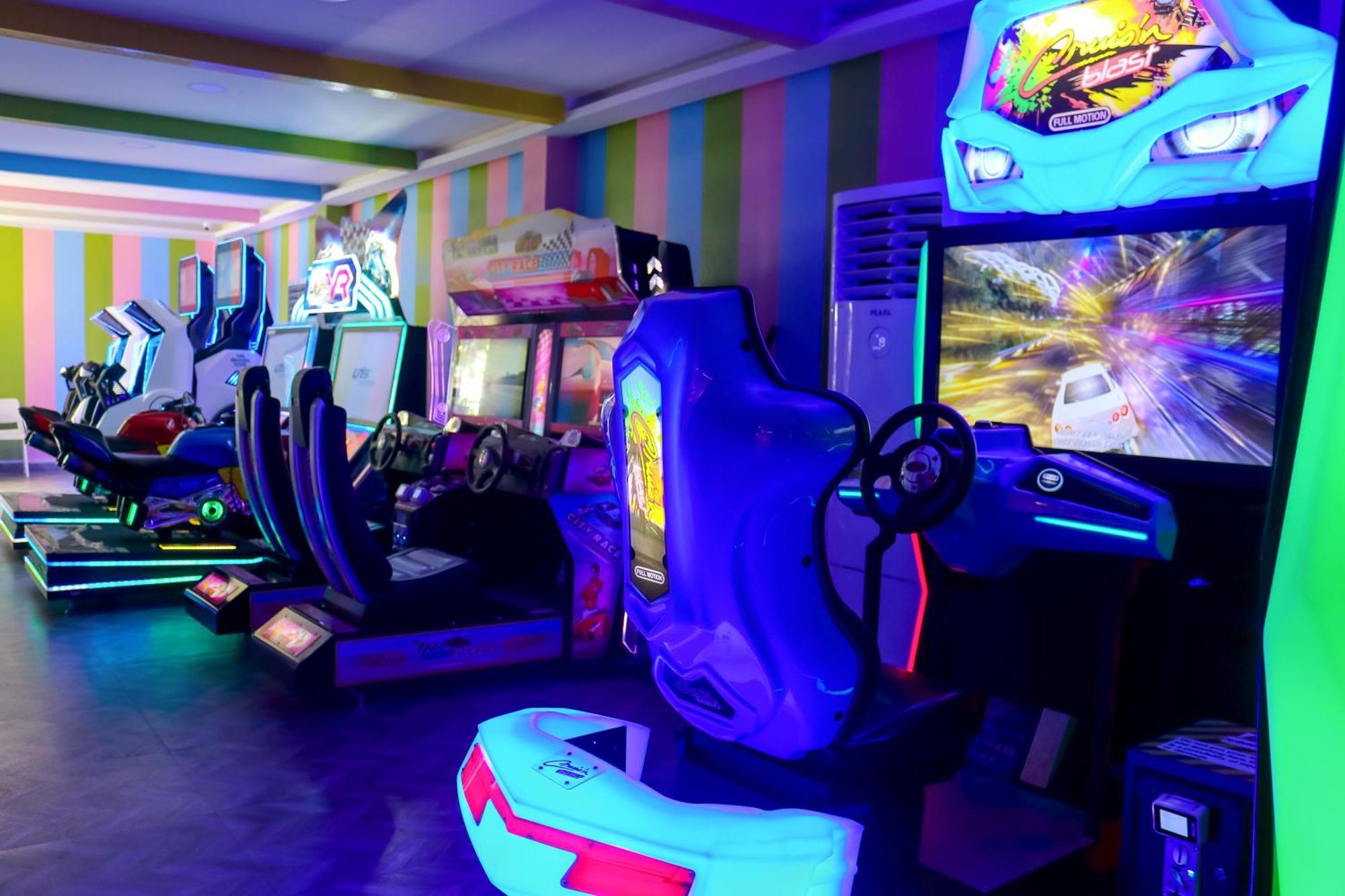 Lagoona Beach Luxury Resort And Spa Manama Exterior photo The photo shows an arcade area filled with various arcade machines. The setting is illuminated with colorful neon lights, creating a vibrant atmosphere. There are several racing game machines with steering wheels and seats, each accompanied by screen