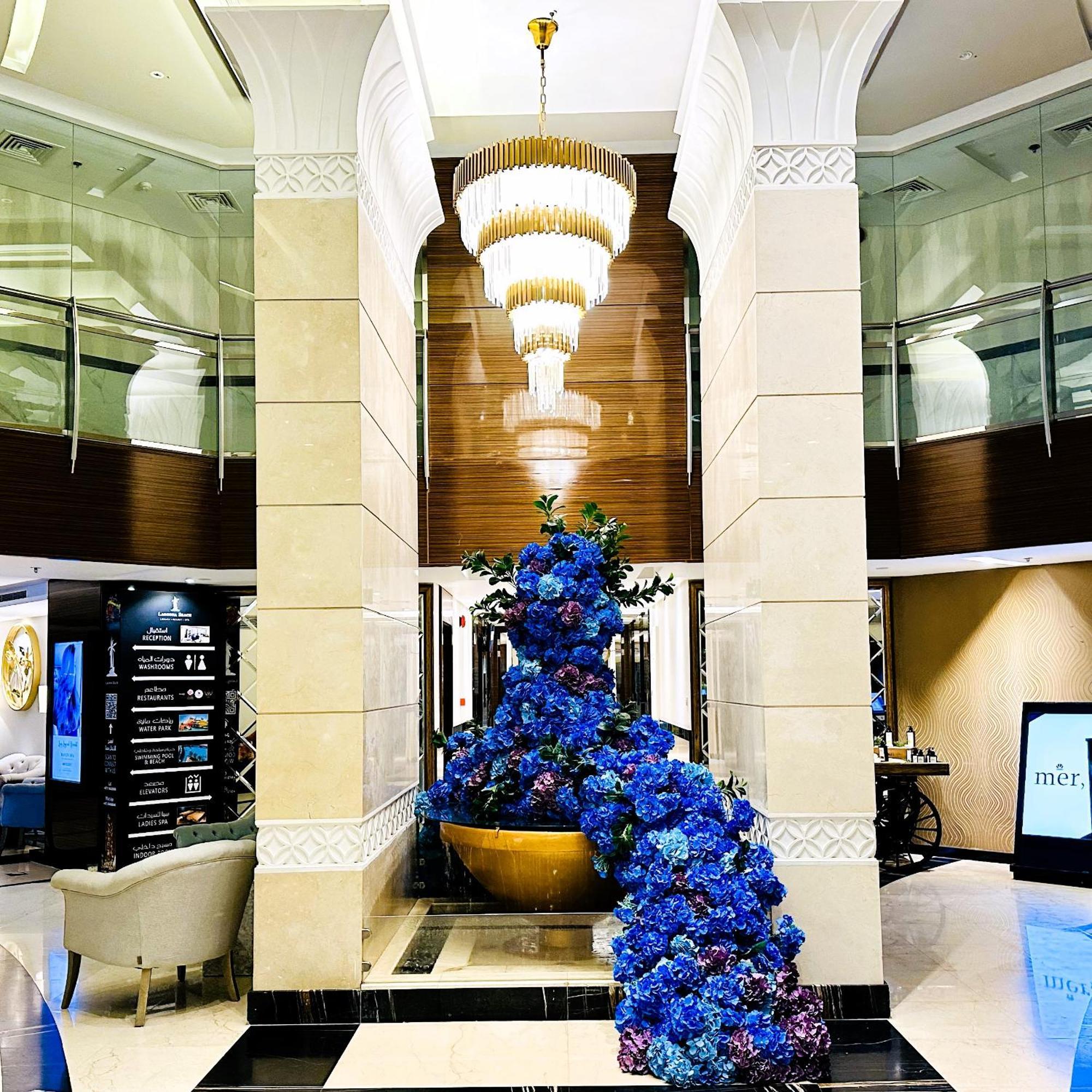 Lagoona Beach Luxury Resort And Spa Manama Exterior photo The photo depicts an elegant lobby or entrance area of a building. Central to the scene is a large floral arrangement featuring vibrant blue flowers. This arrangement is positioned in a decorative bowl. Above it hangs a stylish chandelier with a laye