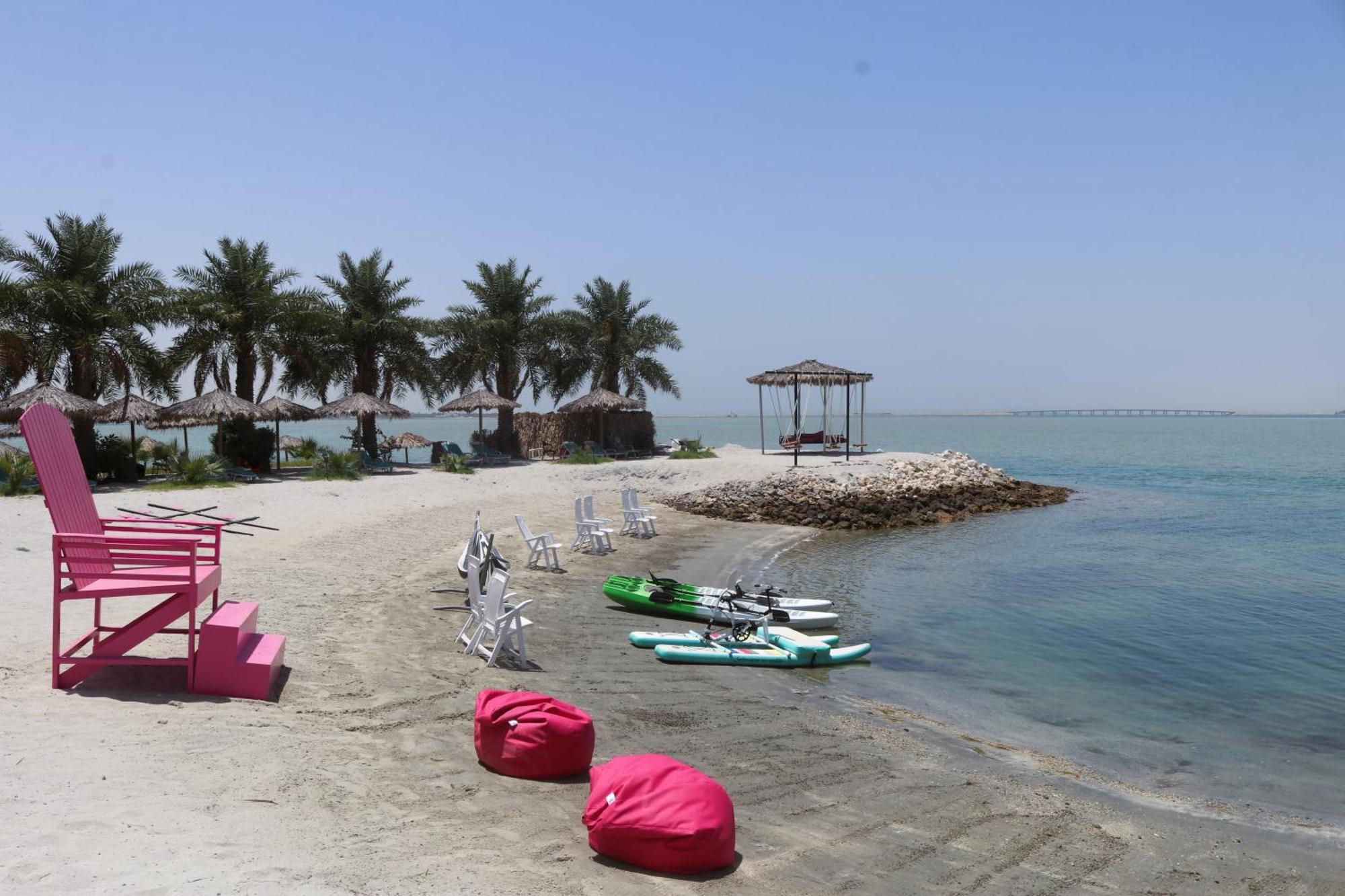 Lagoona Beach Luxury Resort And Spa Manama Exterior photo The photo depicts a serene beach scene with clear blue water and a sunny sky. In the foreground, there are colorful pink bean bags and a large pink chair on the sand. A few white chairs and loungers are positioned near the water's edge. To the side, 