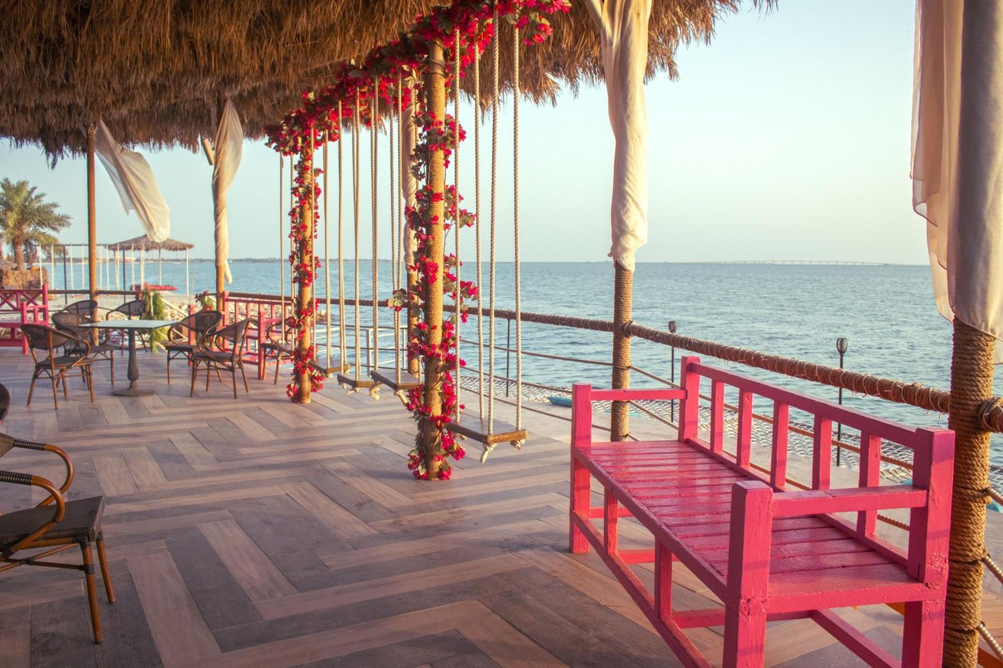 Lagoona Beach Luxury Resort And Spa Manama Exterior photo The photo depicts a scenic outdoor area by the water, with a wooden deck and a thatched roof. Two swings adorned with flowers are hanging from the ceiling, providing a charming touch to the space. In the foreground, there is a bright pink bench, addi