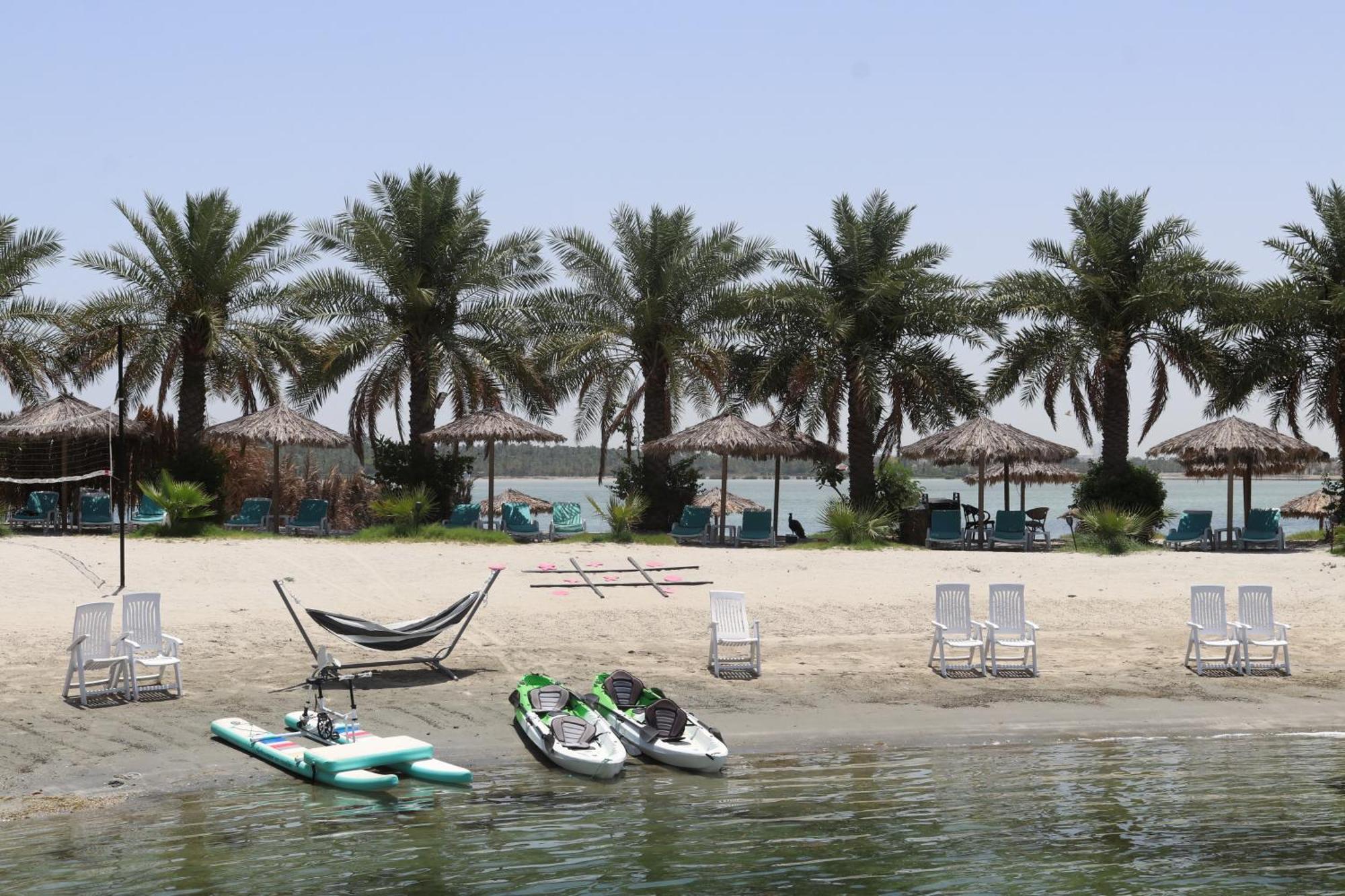 Lagoona Beach Luxury Resort And Spa Manama Exterior photo The photo depicts a serene beach scene with several palm trees lining the shore. There are thatched umbrellas providing shade, and a few lounge chairs are positioned on the sand. A hammock is set up between two supports, inviting relaxation. In the f