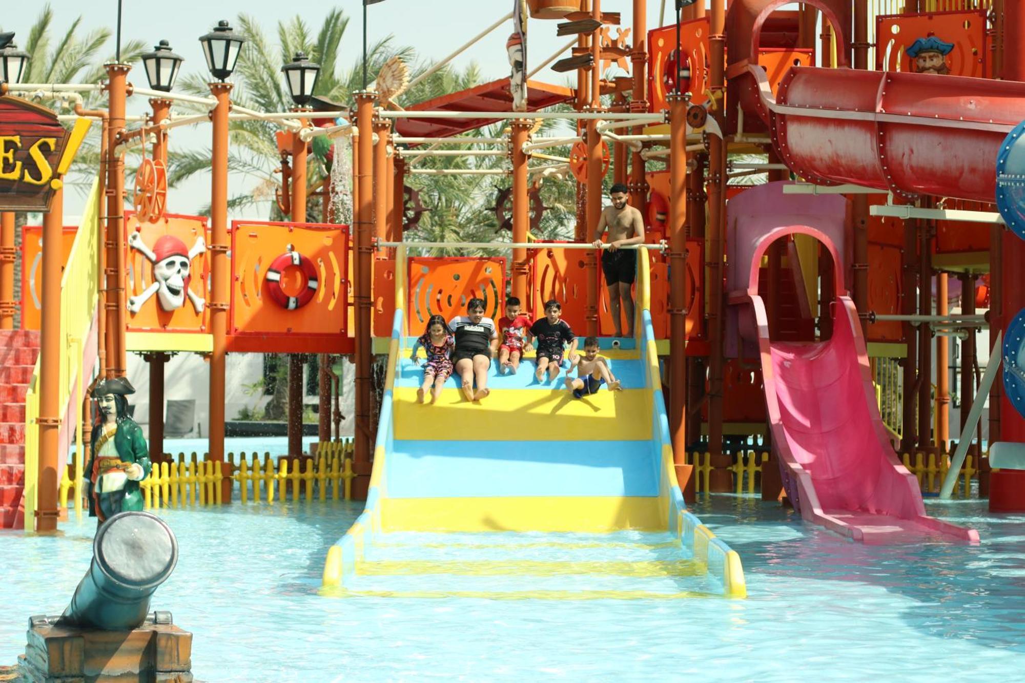 Lagoona Beach Luxury Resort And Spa Manama Exterior photo The photo shows a colorful water park scene featuring a play area with multiple slides. There are children enjoying themselves, sliding down bright yellow and pink slides into a pool of water. The structure has pirate-themed decorations, including a 