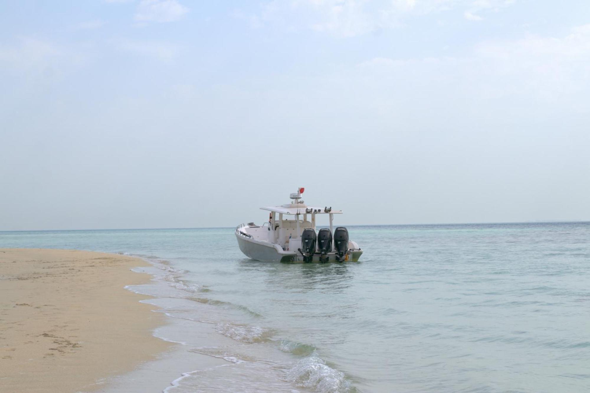 Lagoona Beach Luxury Resort And Spa Manama Exterior photo The photo depicts a small boat anchored near a sandy beach, partially submerged in calm, clear water. The boat is white and features three outboard motors on its stern. In the background, the sea stretches out toward the horizon under a light, cloudy