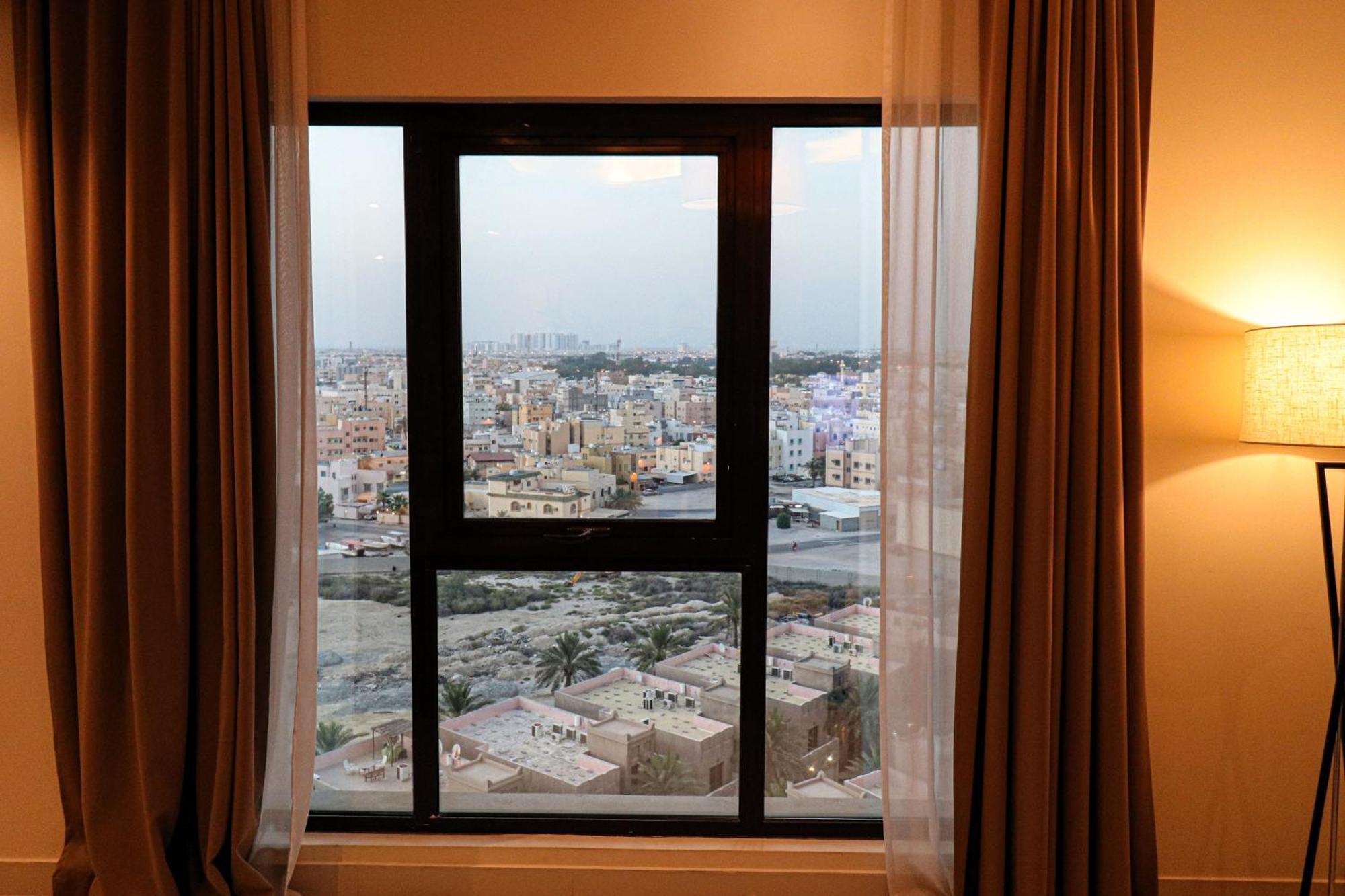 Lagoona Beach Luxury Resort And Spa Manama Exterior photo The photo shows a window with a black frame and sheer curtains. Through the window, there is a view of a cityscape, displaying a densely populated area with various buildings and rooftops. The sky appears slightly overcast, suggesting a muted lightin