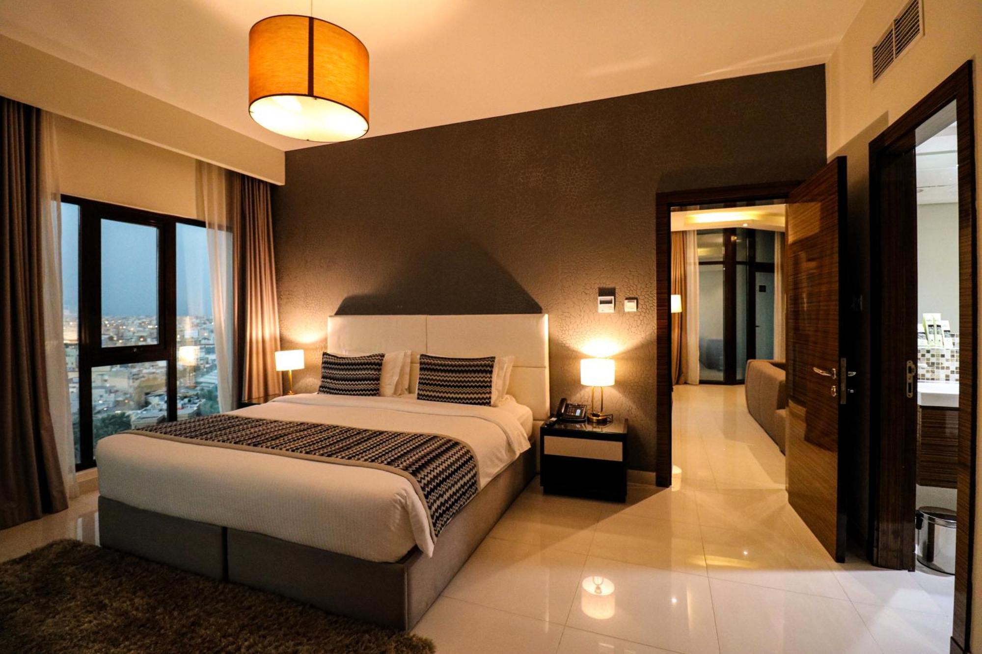 Lagoona Beach Luxury Resort And Spa Manama Exterior photo The photo depicts a modern hotel room or suite. It features a large bed with a white and gray color scheme, accented with decorative pillows. On either side of the bed, there are sleek bedside lamps providing warm lighting. The walls are a dark color