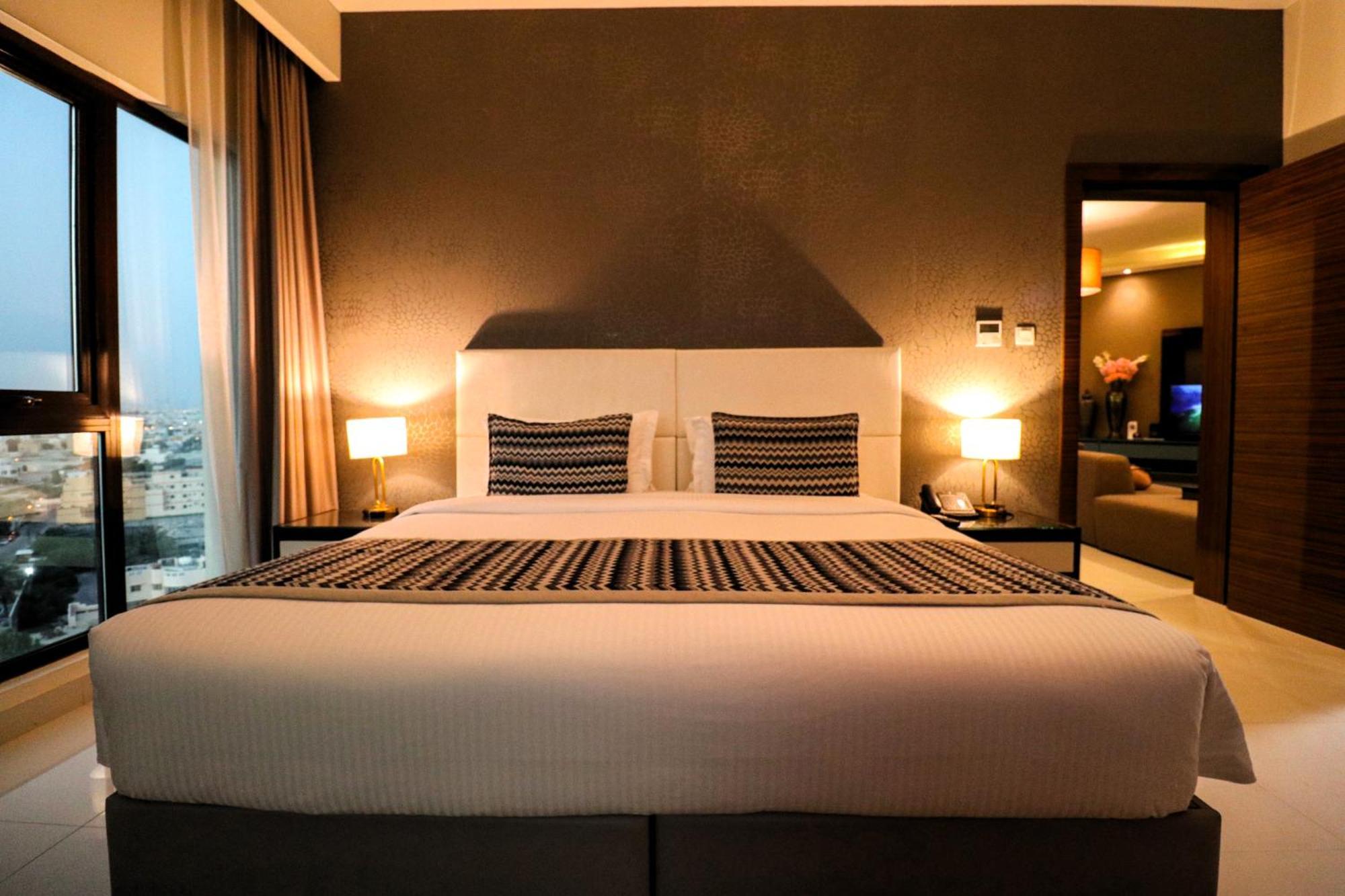 Lagoona Beach Luxury Resort And Spa Manama Exterior photo The photo depicts a modern hotel room featuring a large bed with a white comforter and two decorative pillows. The headboard is light-colored, and the walls are painted a dark hue. On either side of the bed, there are bedside tables with lamps. The r