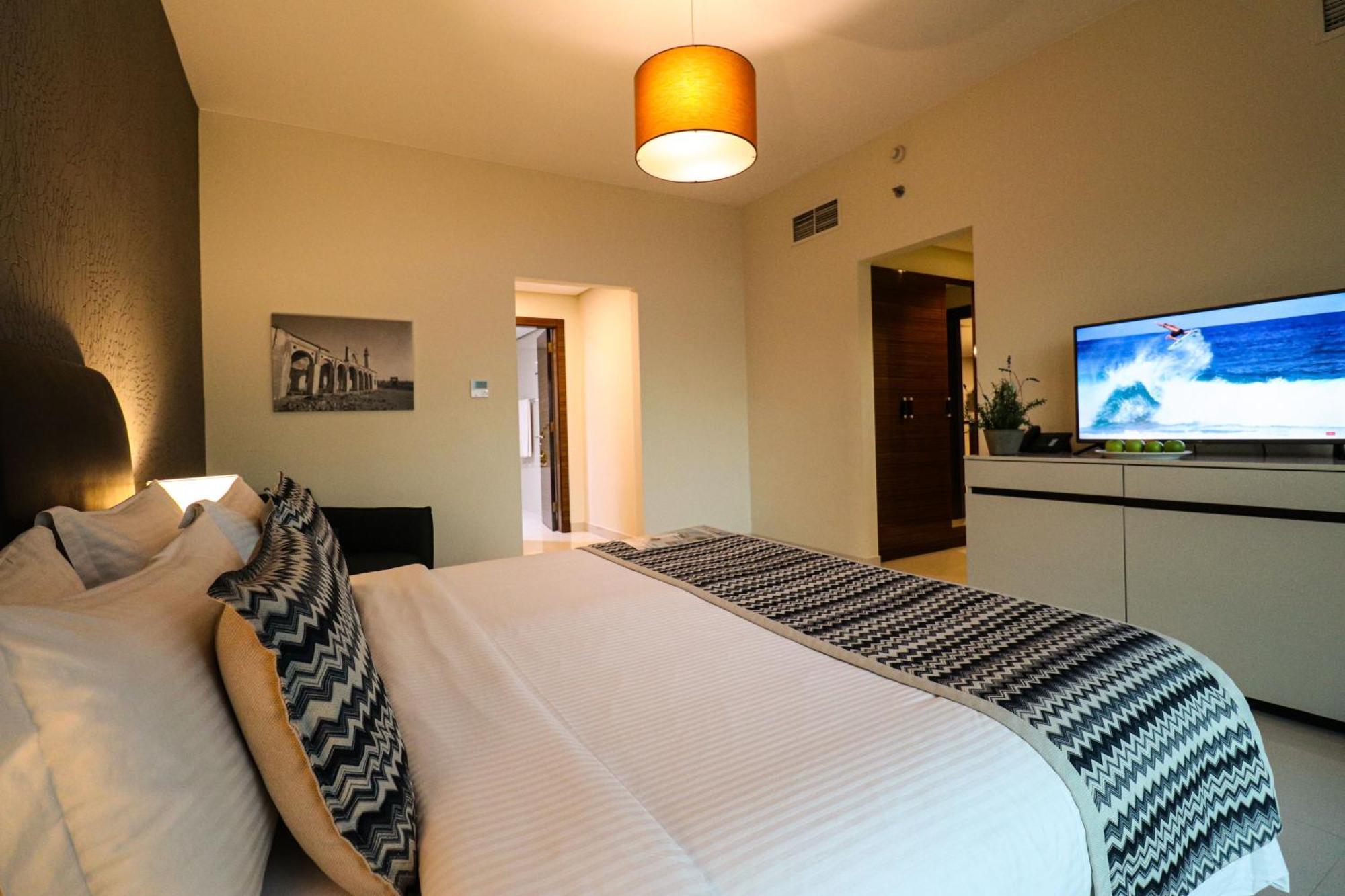 Lagoona Beach Luxury Resort And Spa Manama Exterior photo The photo shows a well-decorated hotel room. There is a large bed with white linens and decorative pillows featuring a zigzag pattern. On the wall beside the bed is a piece of artwork, and there is a modern television on a white furniture unit opposi
