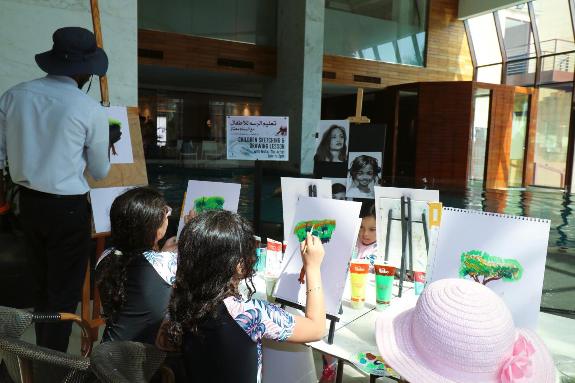 Lagoona Beach Luxury Resort And Spa Manama Exterior photo The photo shows a group of children and an adult engaged in an art activity near a pool area. The children are seated at a table, painting on canvases or paper, with a focus on trees, as indicated by the green paint they are using. An adult, wearing 