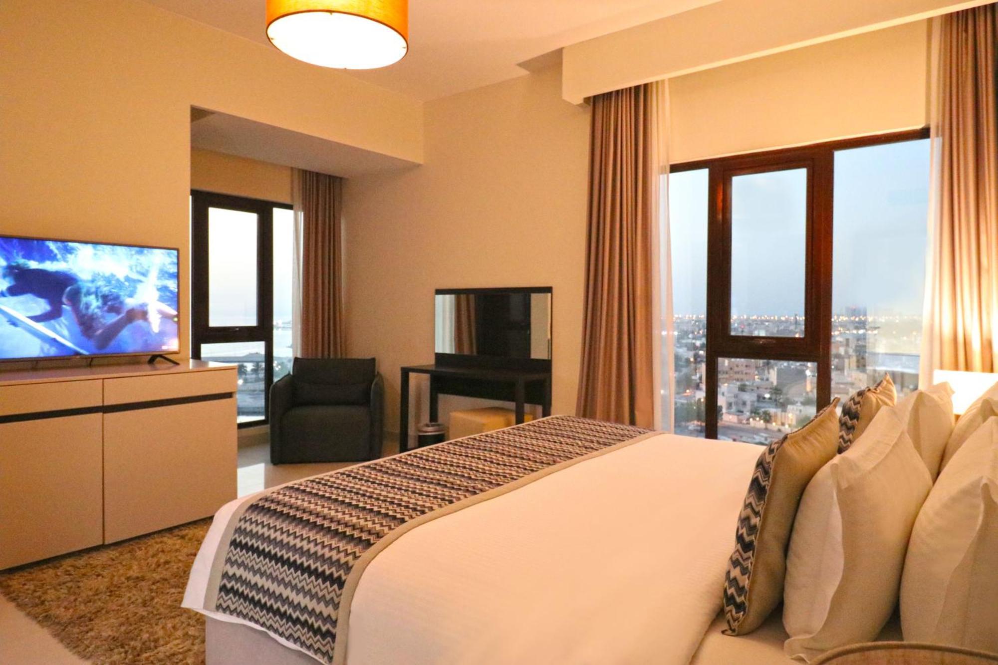 Lagoona Beach Luxury Resort And Spa Manama Exterior photo The photo shows a modern hotel room or suite. It features a neatly made bed with decorative pillows in the foreground. To the left, there is a TV on a cabinet, and further back, a comfortable chair is situated in front of a large window that likely o