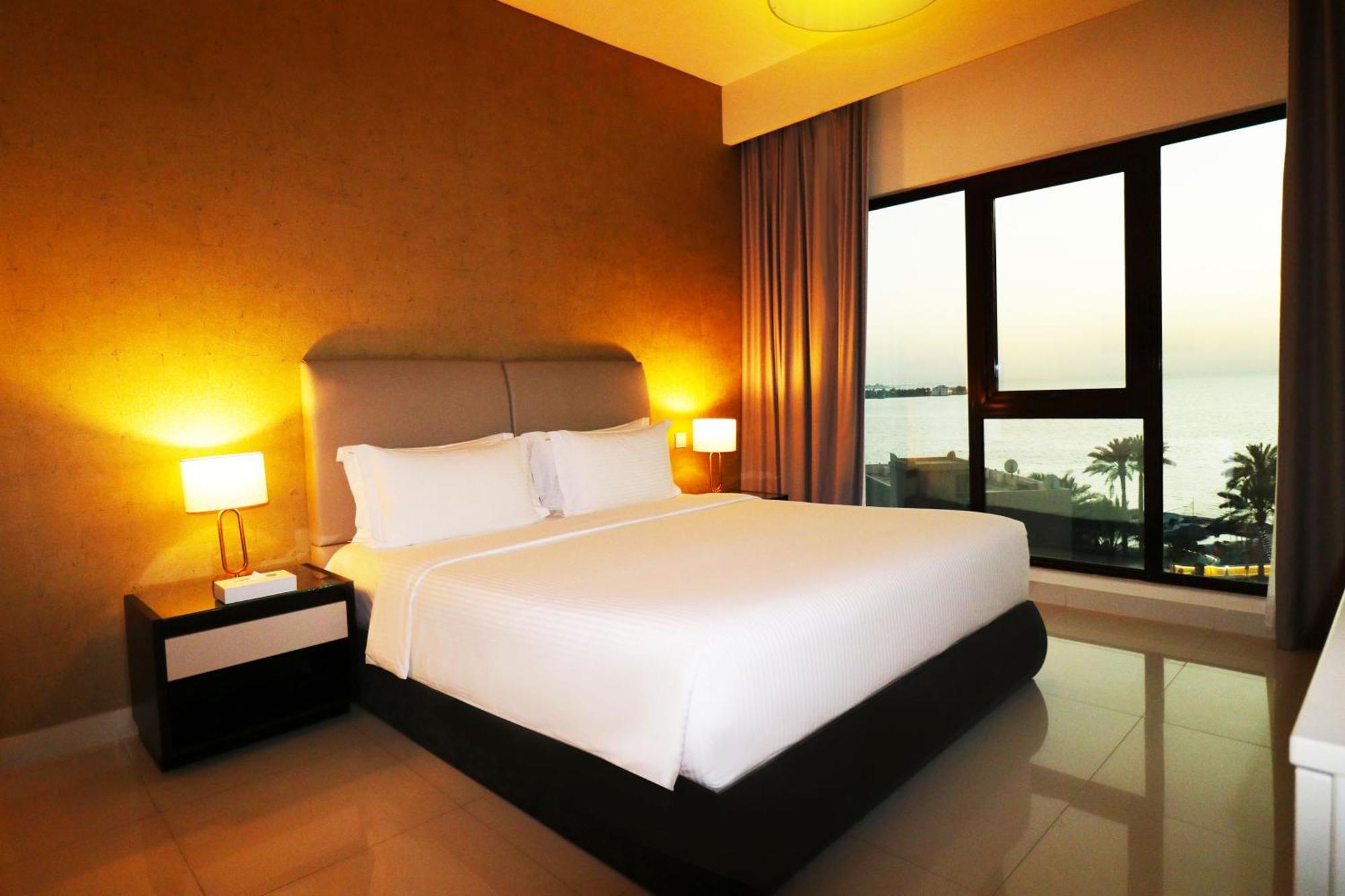 Lagoona Beach Luxury Resort And Spa Manama Exterior photo The photo shows a modern hotel bedroom with a large, comfortable bed dressed in white linens. The room features warm lighting, including bedside lamps, creating a cozy atmosphere. There are large windows with a view of the outdoors, possibly overlook